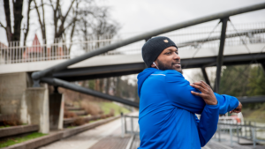 African American man wearing a blue jacket and a hat stretching before exercising outside by trees and a bridge.